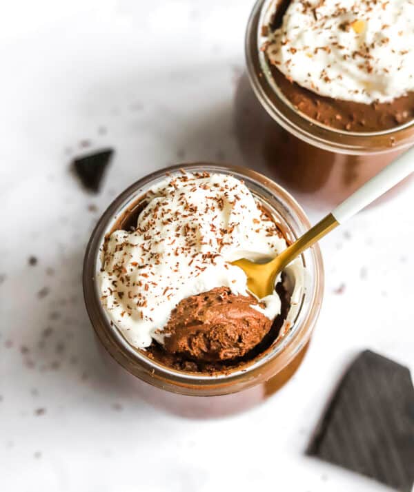Overhead view of a jar filled with silky chocolate mousse with whipped cream on top of it with a spoon in the jar and another jar of mousse behind it.