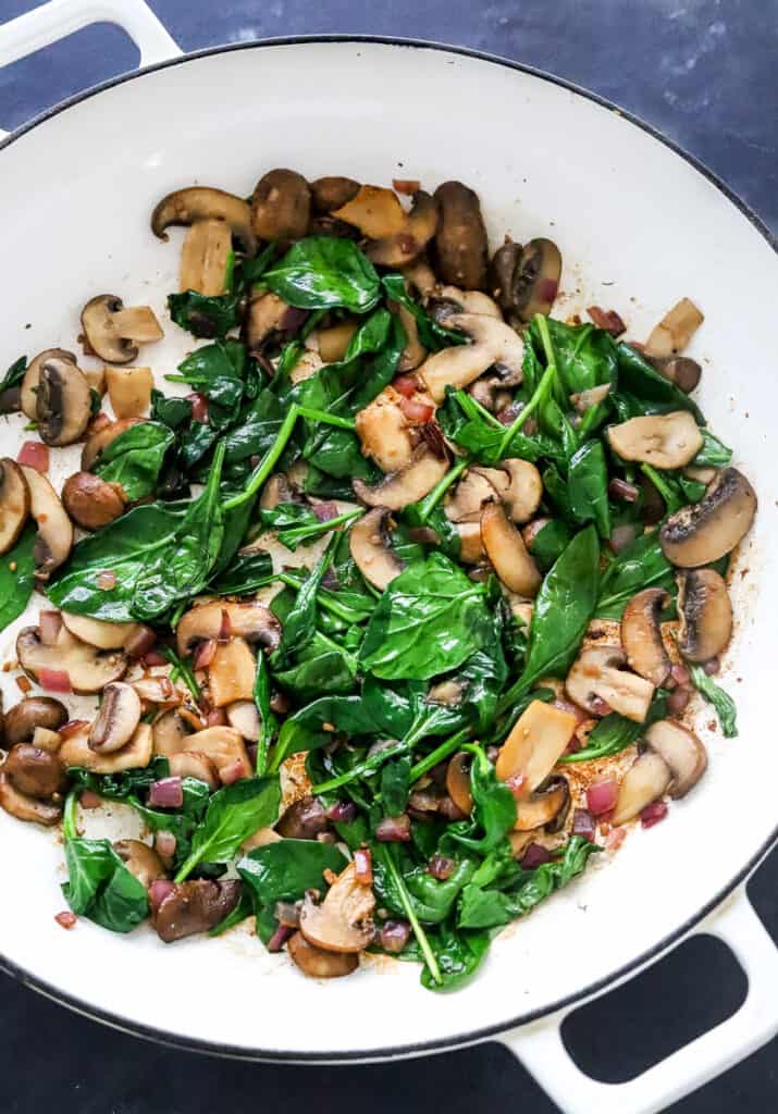 Cooked spinach, mushrooms and red onion in a round white pan. 