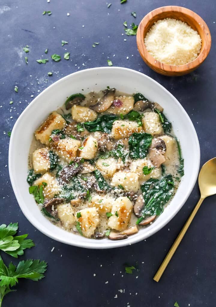 Cauliflower gnocchi in a bowl mixed in a creamy mushroom spinach sauce with a spoon next to it and a bowl of parmesan cheese behind it. 