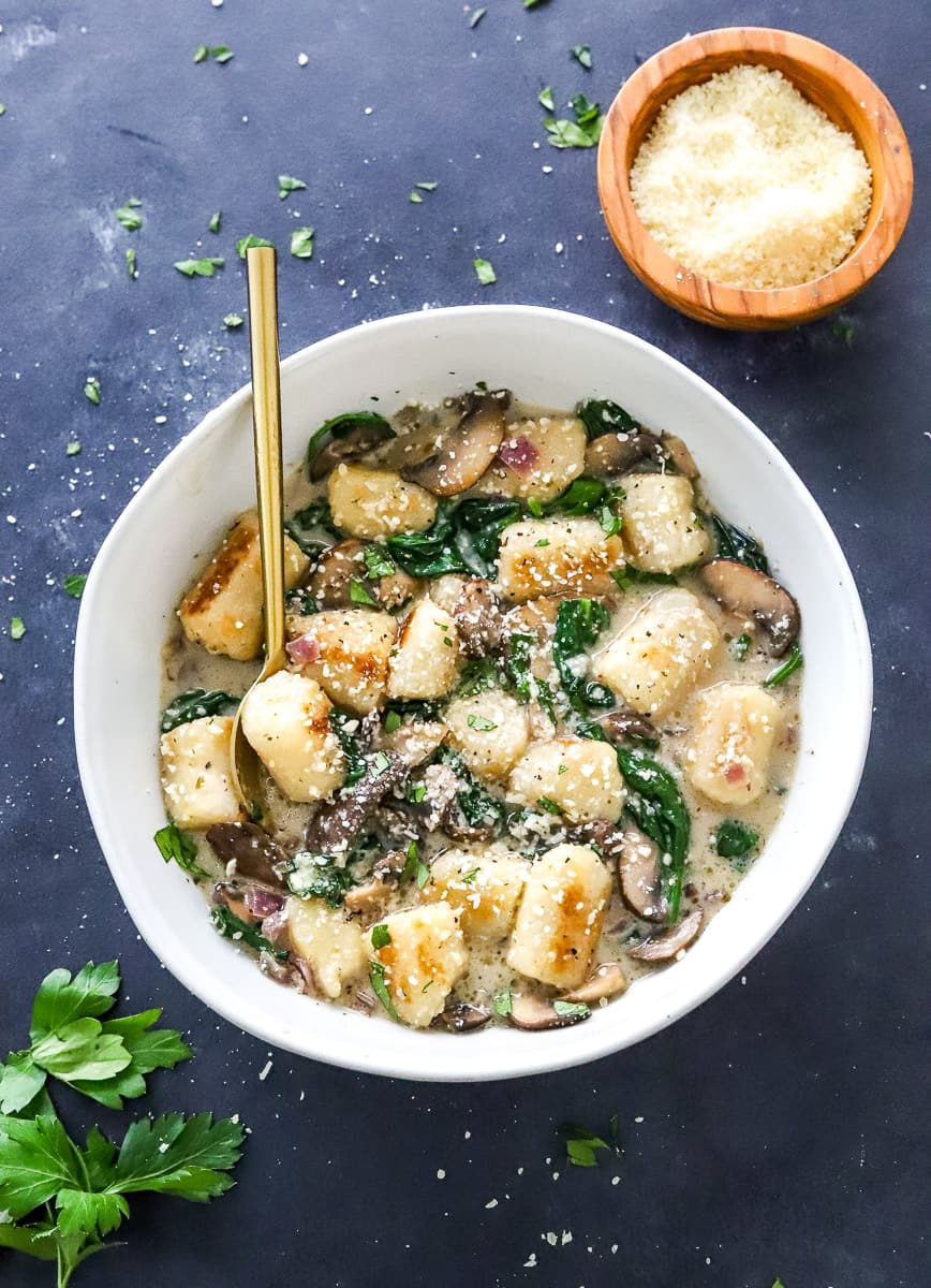 Bowl of trader joe's cauliflower gnocchi in a creamy mushroom sauce with spinach with a gold spoon in the bowl. With a bowl of grated parmesan and bunch of green parsley in front of it. 