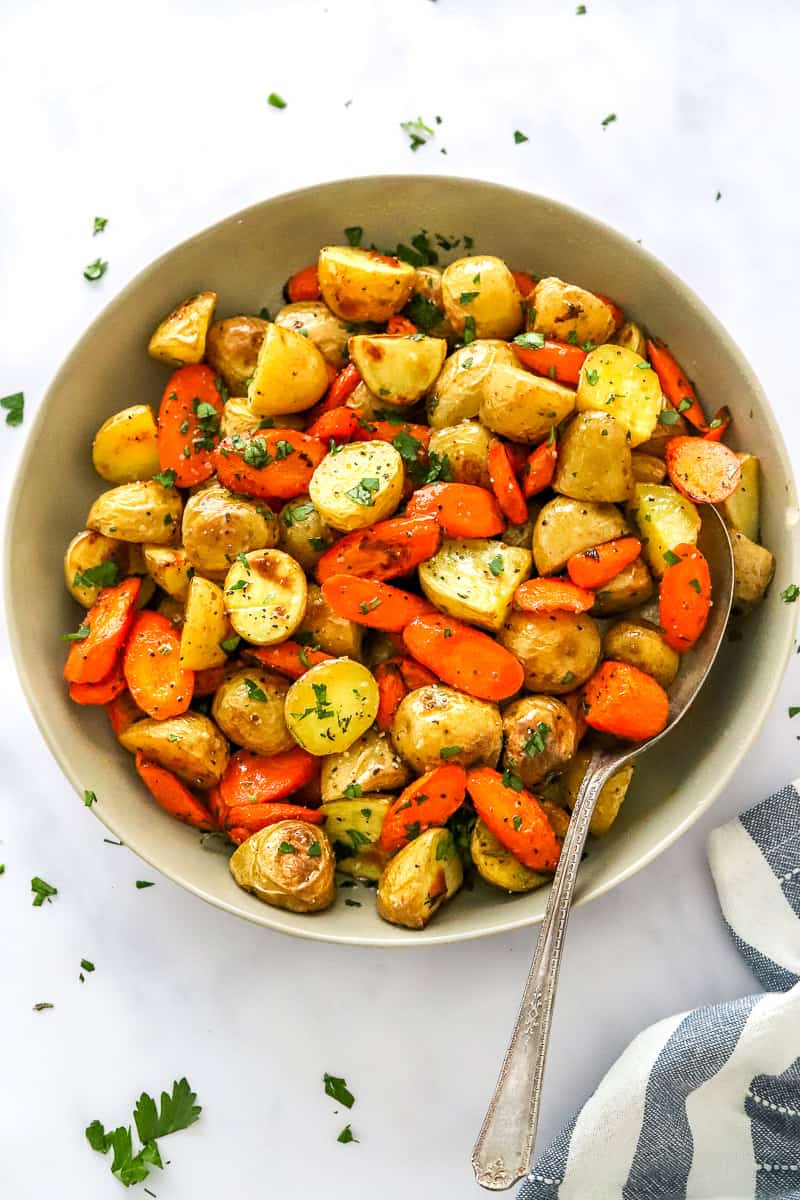 Tan bowl filled with cooked baby potatoes and carrots topped with green herbs with a silver serving spoon in the dish. 