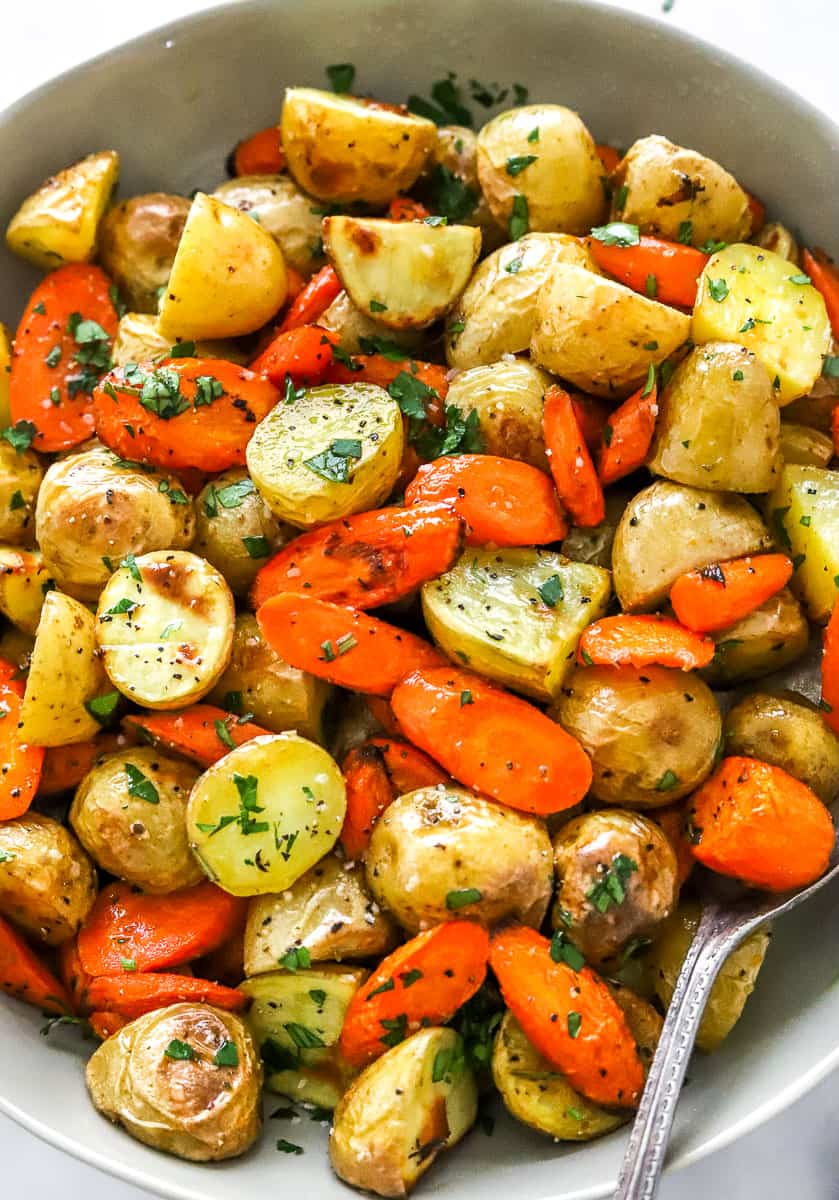 Golden crispy roasted potatoes and carrots in a tan bowl topped with green herbs with a spoon in the bowl. 