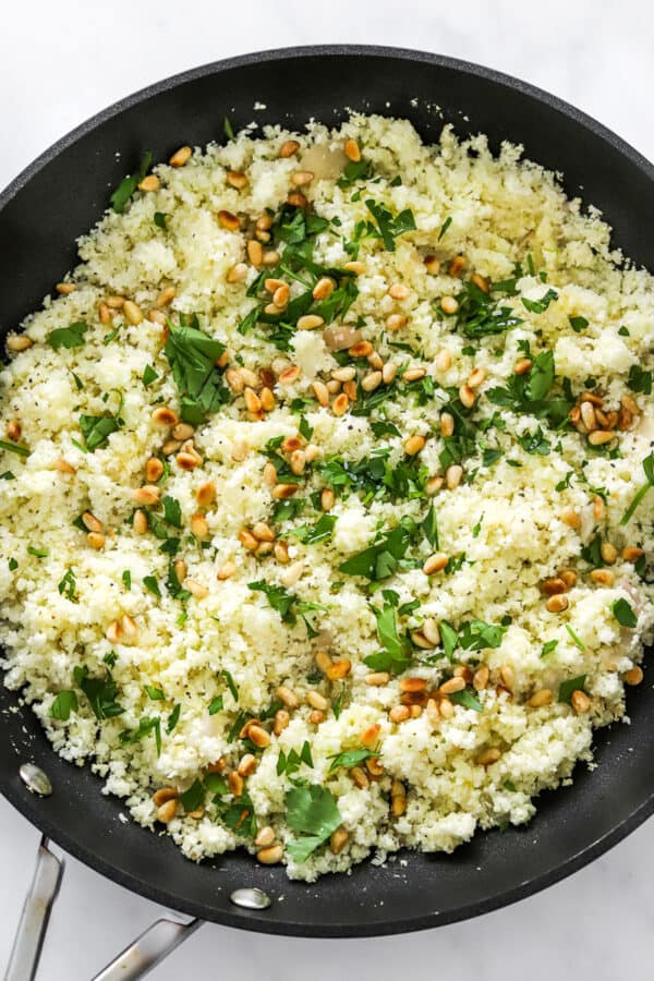 Cooked cauliflower rice topped with parsley and pine nuts in a skillet.