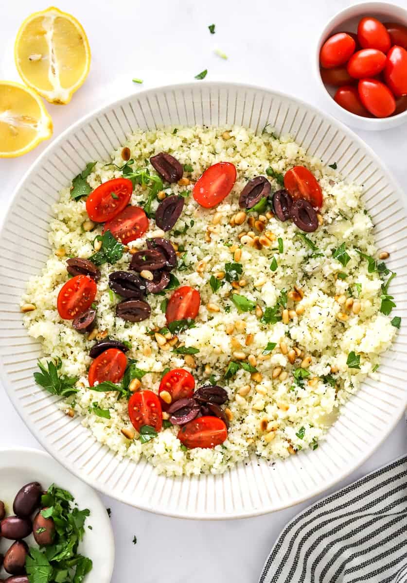 Mediterranean cauliflower rice in a large bowl topped with sliced tomatoes, olives, herbs, and pine nuts with more tomato and sliced lemon behind it and a striped towel and plate of olive and herbs in front of it.
