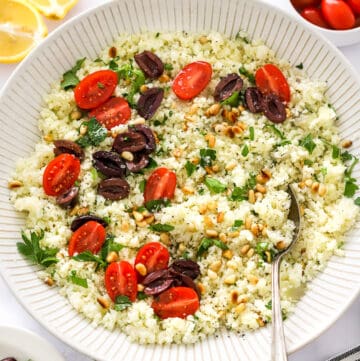 Large bowl filled with cooked cauliflower rice with veggies on top of it with a spoon in the bowl and more tomatoes and sliced lemon behind it.