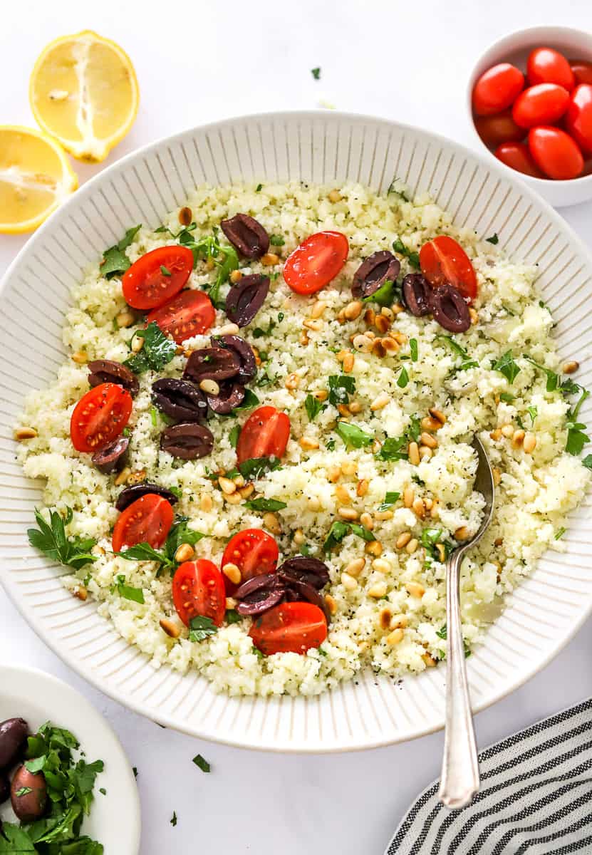 Large bowl filled with cooked cauliflower rice with veggies on top of it with a spoon in the bowl and more tomatoes and sliced lemon behind it.