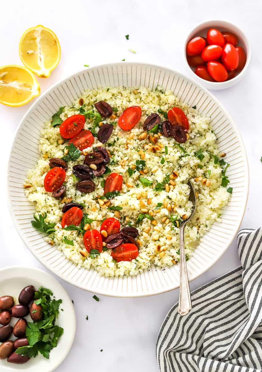 Riced, cooked cauliflower in a large serving bowl topped with sliced tomatoes, Calmat olives, parsley, and pine nuts with a spoon in the bowl more tomatoes and lemon behind it and a striped linen and olives on a plate in front of if.