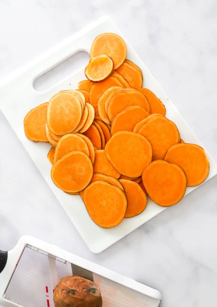 Thinly sliced sweet potato rounds on a white cutting board with a mandolin sliced with a sweet potato in it in front of it. 
