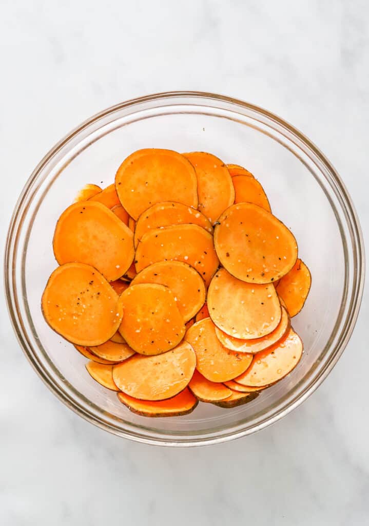 Sliced , raw sweet potatoes in a bowl seasoned with oil, salt and pepper. 