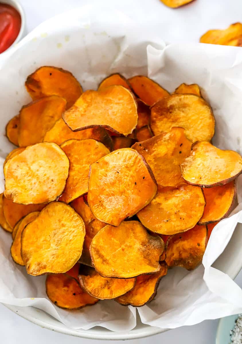 Air fryer sweet potato chips in a white parchment paper covered basket with a bowl of ketchup behind it.