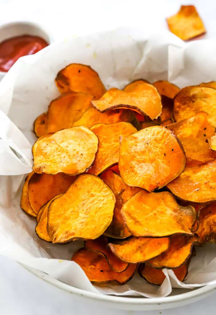 A pile of crispy, sweet potato chips in a basket on white paper with a small white bowl of ketchup behind it. 