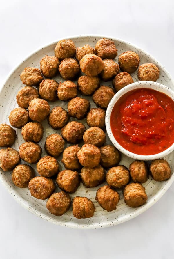 Round white plate filled with cooked brown meatballs with a white bowl filled with tomato sauce next to them on the plate.
