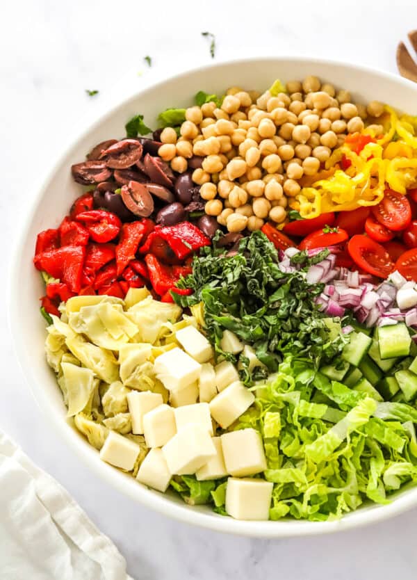 Chopped lettuce, cheese, peppers, olives, onions, and tomatoes in a large salad bowl with a white linen in front of them.