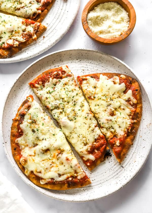 Plate of cooked naan pizza sliced into 3 slices with another plate of the pizza and a bowl of grated parmesan behind it.