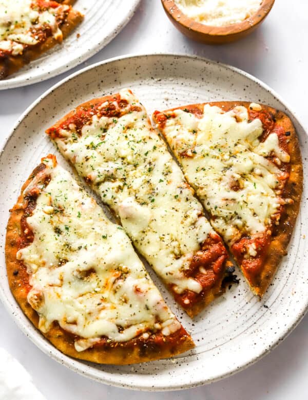 Baked naan pizza with melted cheese and red sauce on top of it cut into 3 pieces with another plate with more naan pizza and a brown bowl of grated cheese behind it.