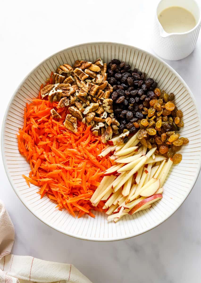 Bowl filled with shredded orange carrots, sliced apples, gold and black raisins, and chopped pecans with a jar of white dressing behind it and a linen in front of it. 