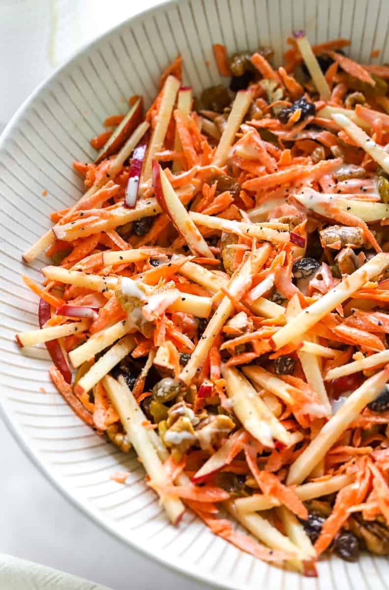 Close up of tossed shredded carrot, apple salad in a bowl covered in a creamy dressing with raisins and chopped nuts in it. 