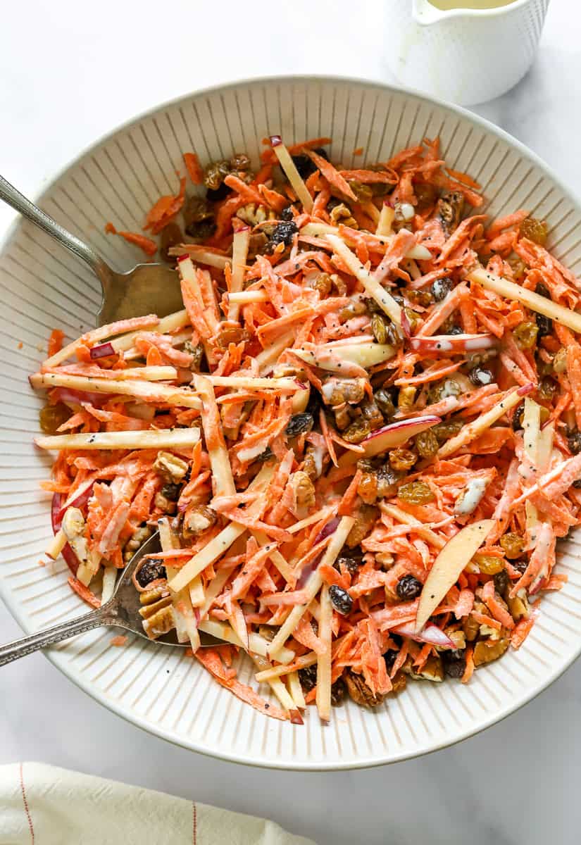 Shredded carrot raisin salad in a round salad bowl tossed in a creamy dressing with silver salad tongs in the bowl. 