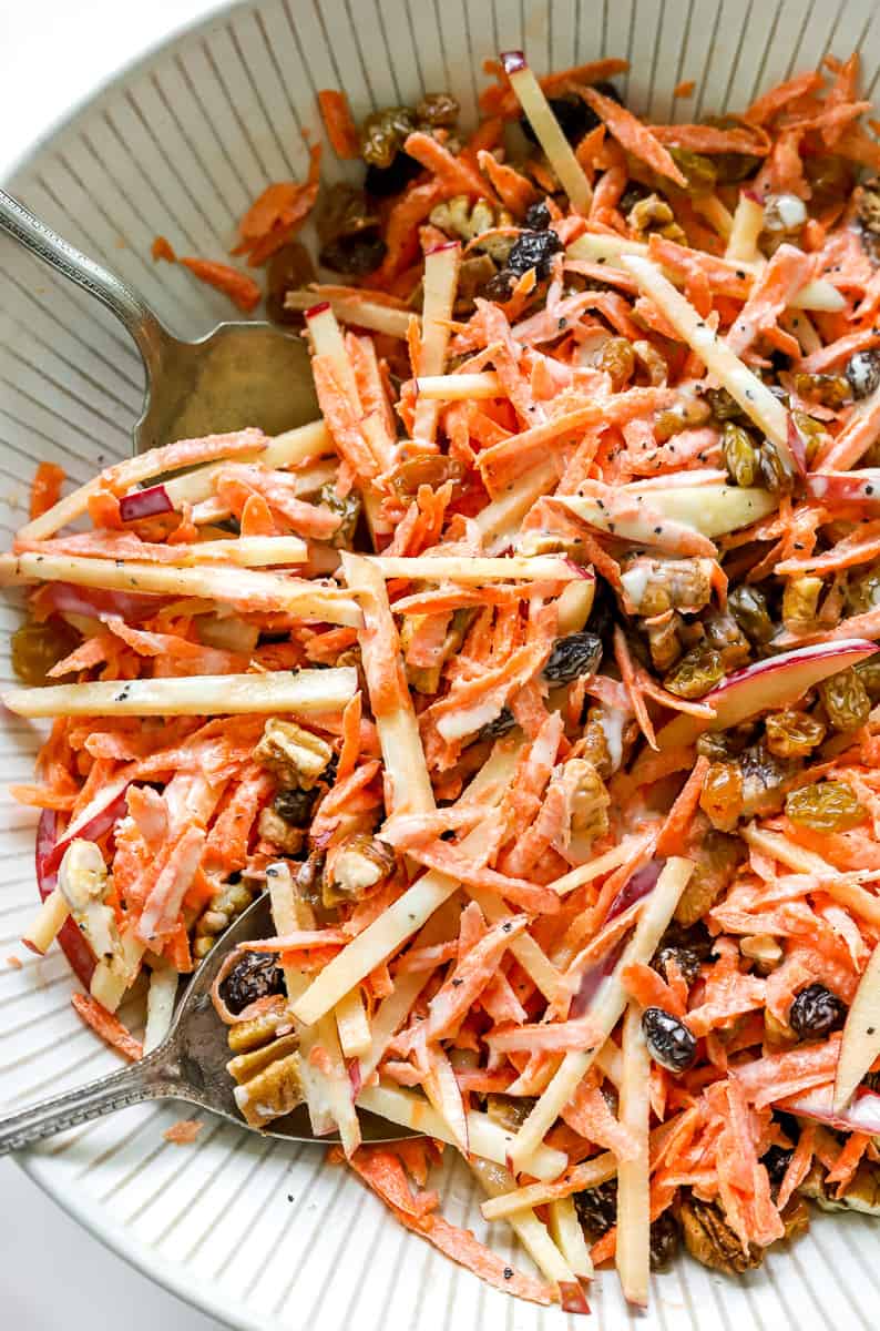 Close up of shredded carrots, sliced apples, nuts and raisins covered in a creamy dressing tossed in a bowl with serving spoons in the bowl with the salad. 