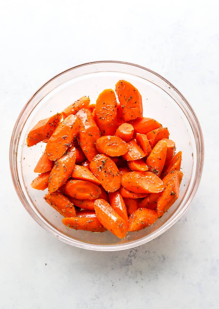 Glass mixing bowl with with raw seasoned, peeled, cut carrots. 