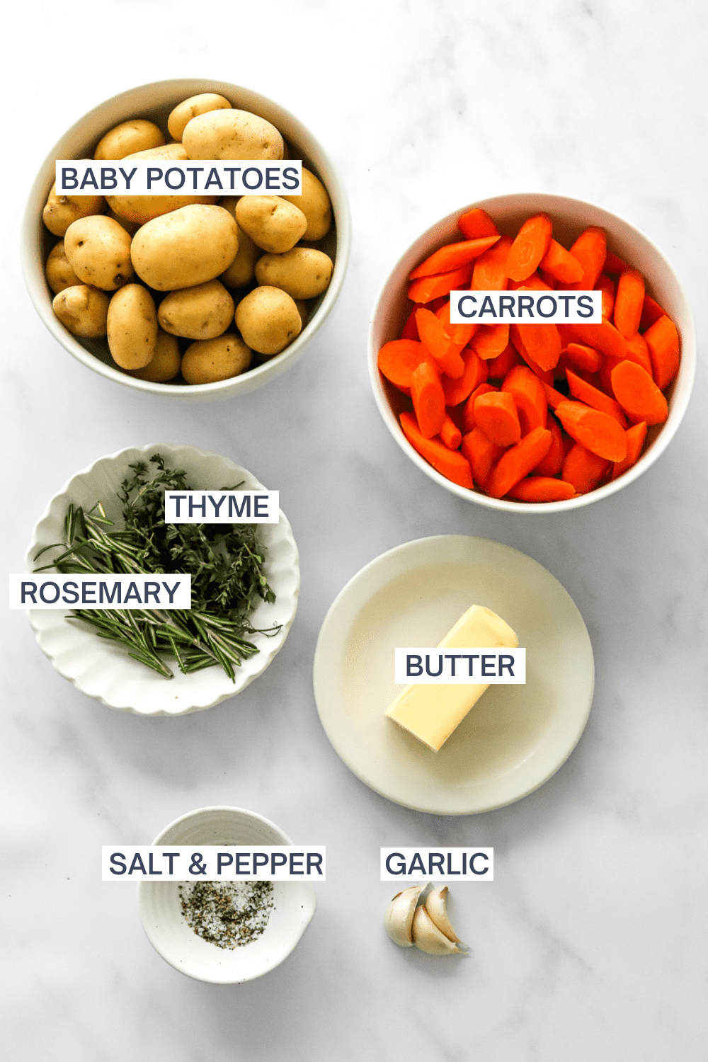 Baby gold potatoes, chopped carrots, herbs, butter, salt and pepper and garlic cloves in bowls with labels over each ingredient. 