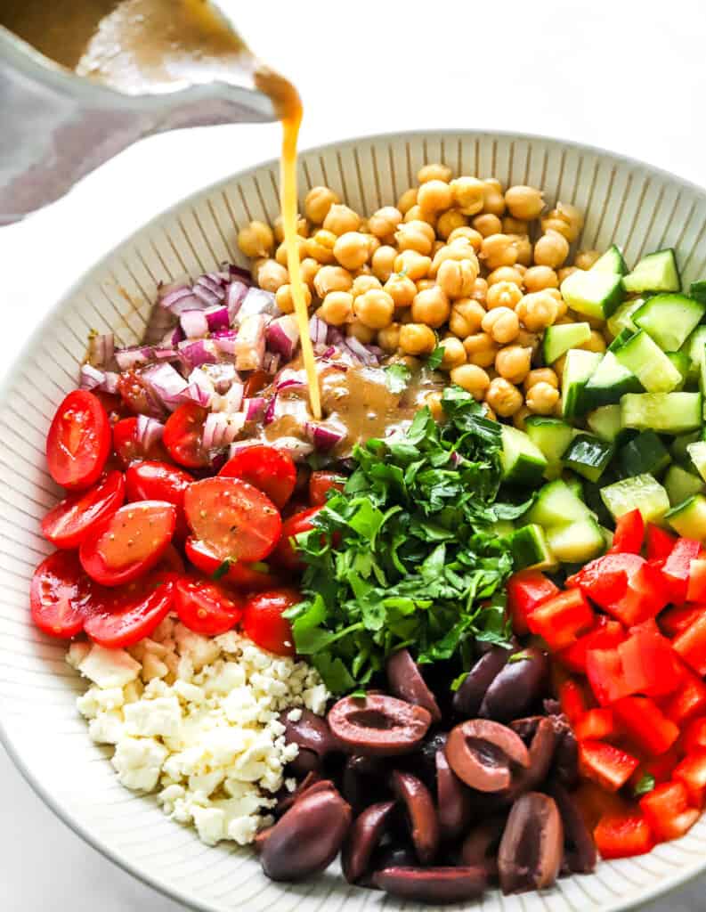 Salad dressing being poured onto a Mediterranean bowl of chickpeas, fresh chopped veggies and herbs. 