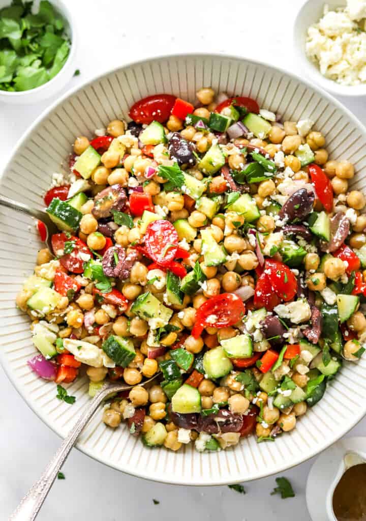 Large salad bowl filled with a chopped Mediterranean Chickpea Salad with serving spoons in the bowl and a bowl of chopped herbs and bowl of crumbled feta cheese behind it. 