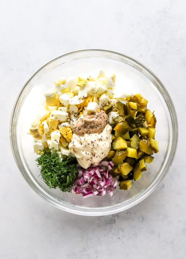 Diced hard boiled eggs, chopped pickles, diced red onion, dill, mayo and mustard in a round glass bowl with salt and pepper sprinkled on top of it.