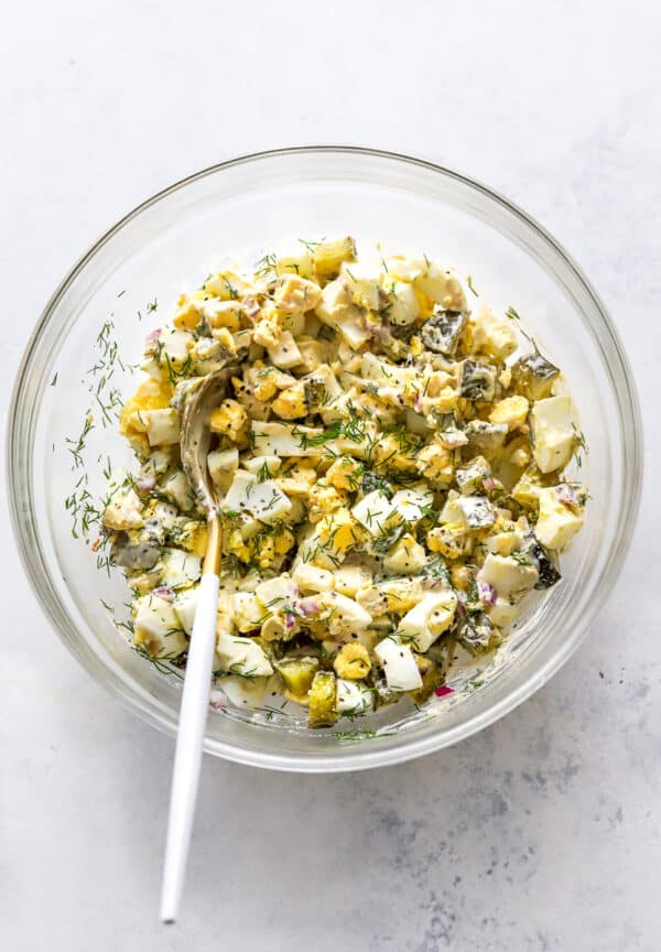 Mixed creamy healthy egg salad in a round glass mixing bowl with a spoon in the bowl.