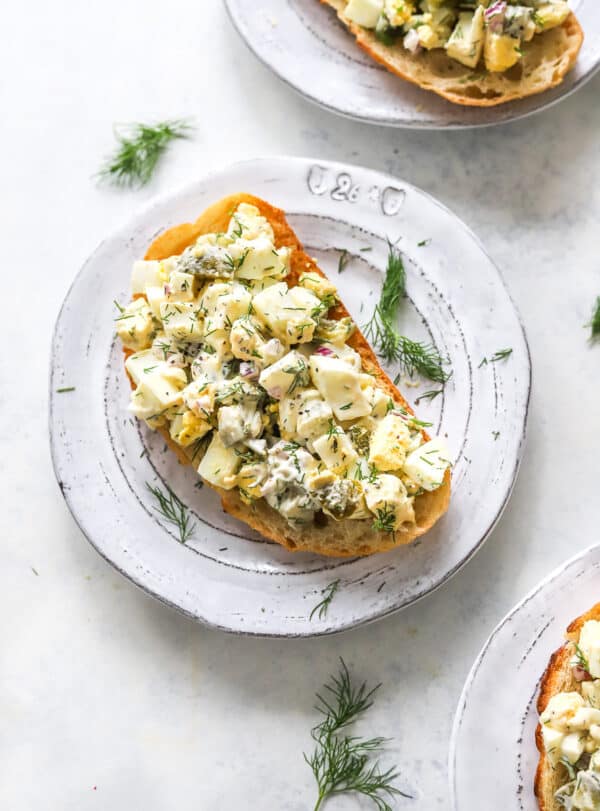 Healthy egg salad on toasted sourdough toast on a round plate with another plate of it behind it and a plate of it in front of it with some fresh herbs around it.