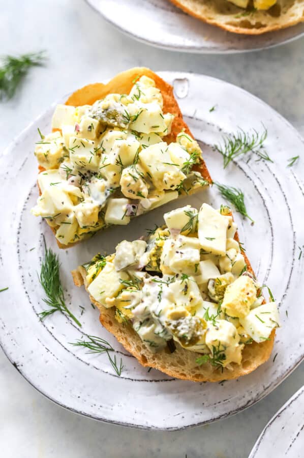Plate with a piece of toast cut in half with creamy egg salad piled on top of it with fresh dill sprinkled around it.