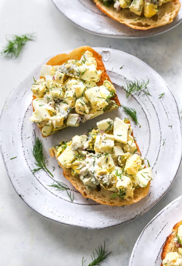 Egg salad on toast cut in half on a plate with another plate of it in front of it and behind it.