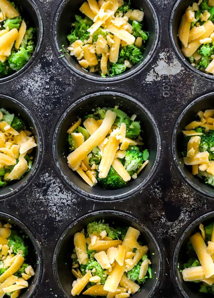 Black muffin pan filled with broccoli and shredded cheddar cheese.