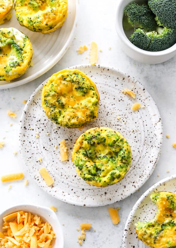 Egg muffins with broccoli and cheese in them on a plate with more egg muffins in front of them and behind them.