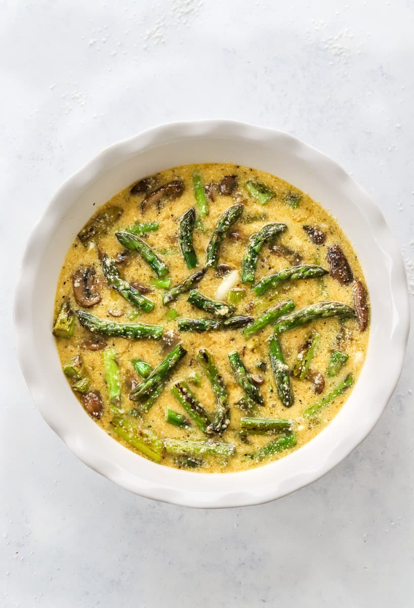 White pie dish filled with whisked eggs and cooked asparagus and mushrooms.
