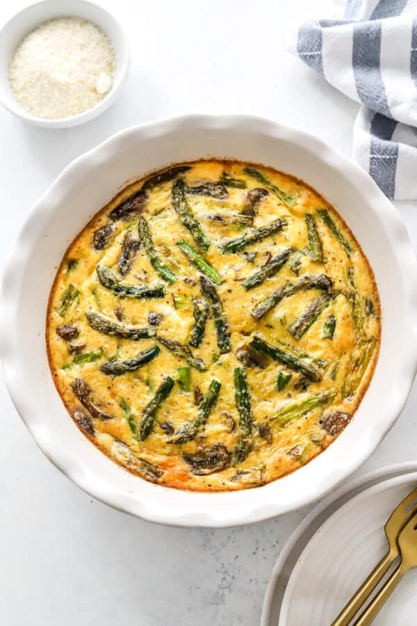 Asparagus crustless quiche in a white pie dish with plates and forks in front of it and a bowl of grated cheese and a striped towel behind it.