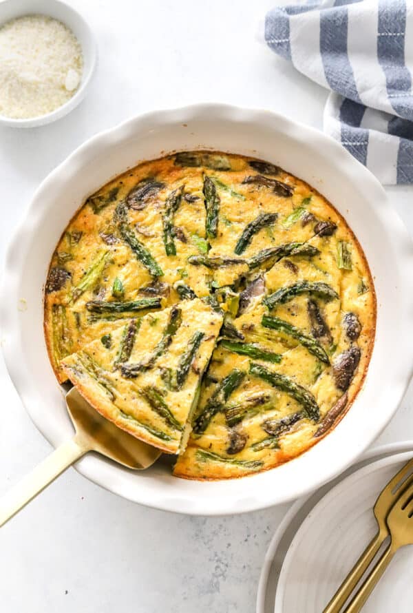 Vegetable quiche on a tables with a sliced being removed from it with plates and gold forks in front of it and a bowl of grate cheese and a striped linen behind it.