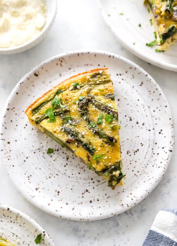 Plate with a sliced of asparagus quiche with another plate of it behind it and in front of it.