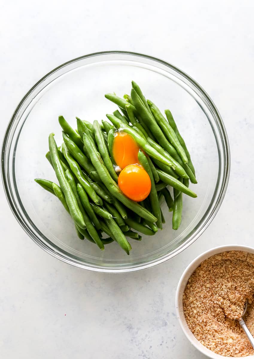 Bowl of uncooked green beans with egg yolks on them with a bowl of breading in a bowl in front of it.