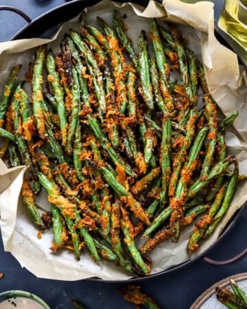 Pile of crispy golden green bean fries in a round platter with a bowl of dipping sauce and more green beans in front of it.