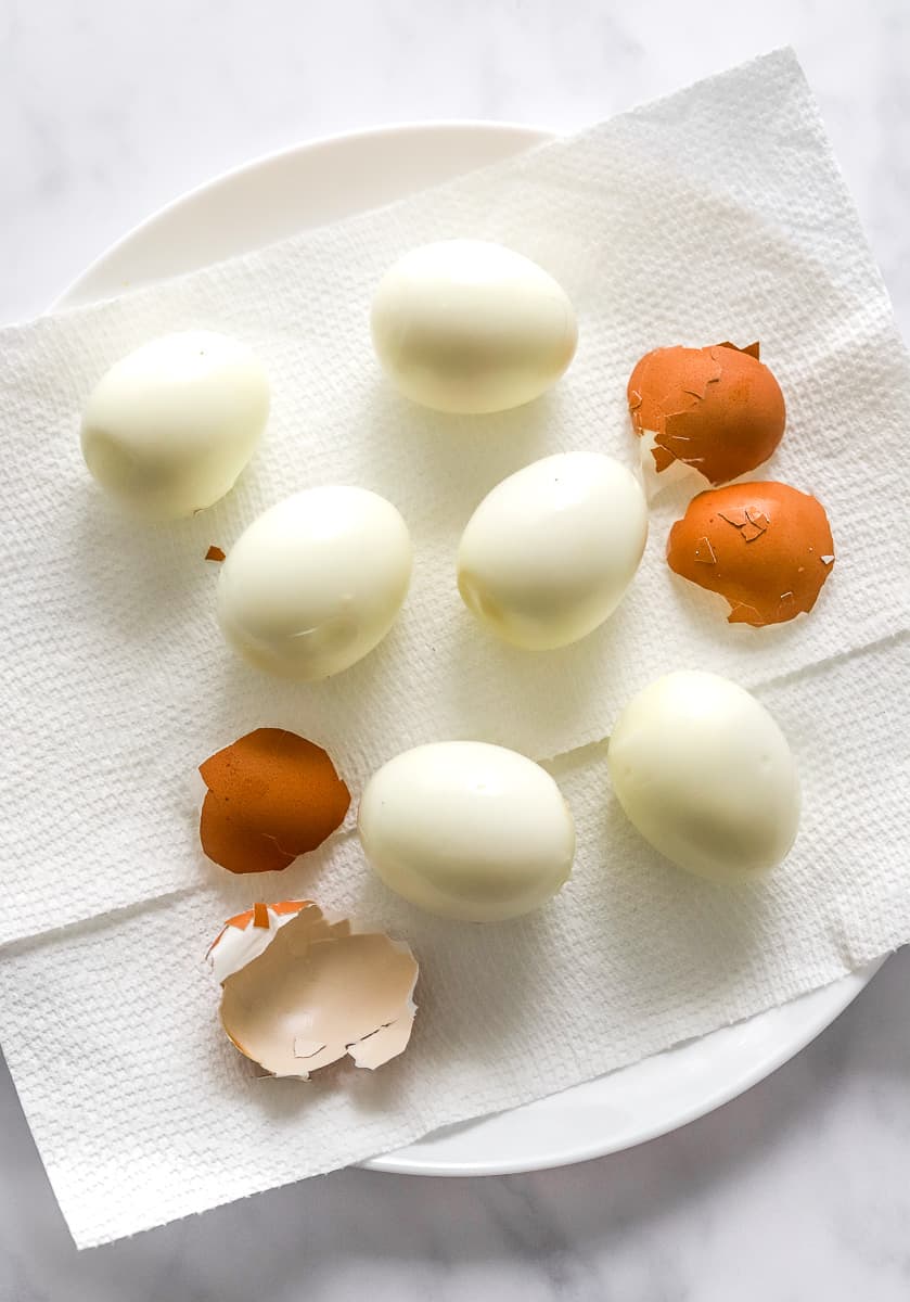 Peeled whole hard boiled eggs on a plated lined with a white paper towel with a couple of egg shells on the plate next to the eggs. 