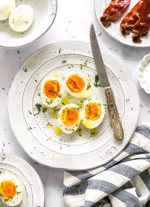 Plate of sliced hard boiled eggs seasoned with herbs with more eggs and bacon behind it and more eggs and a striped linen in front of it.