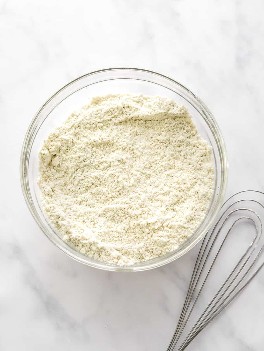 White flour in a glass mixing bowl with a flour whisk next to it.