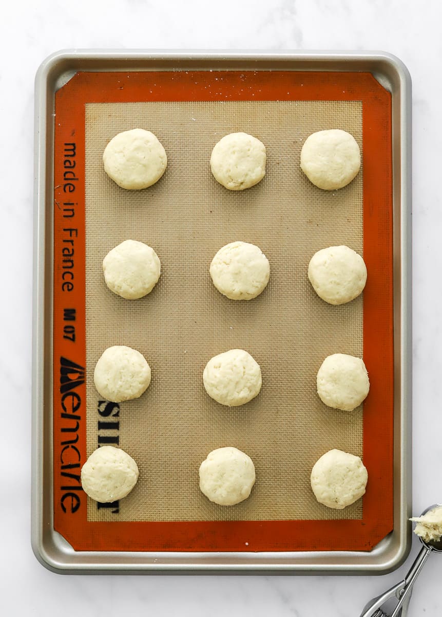 Unbaked round cookies on a baking sheet lined with a baking mat with a scoop with dough in it in front of it.