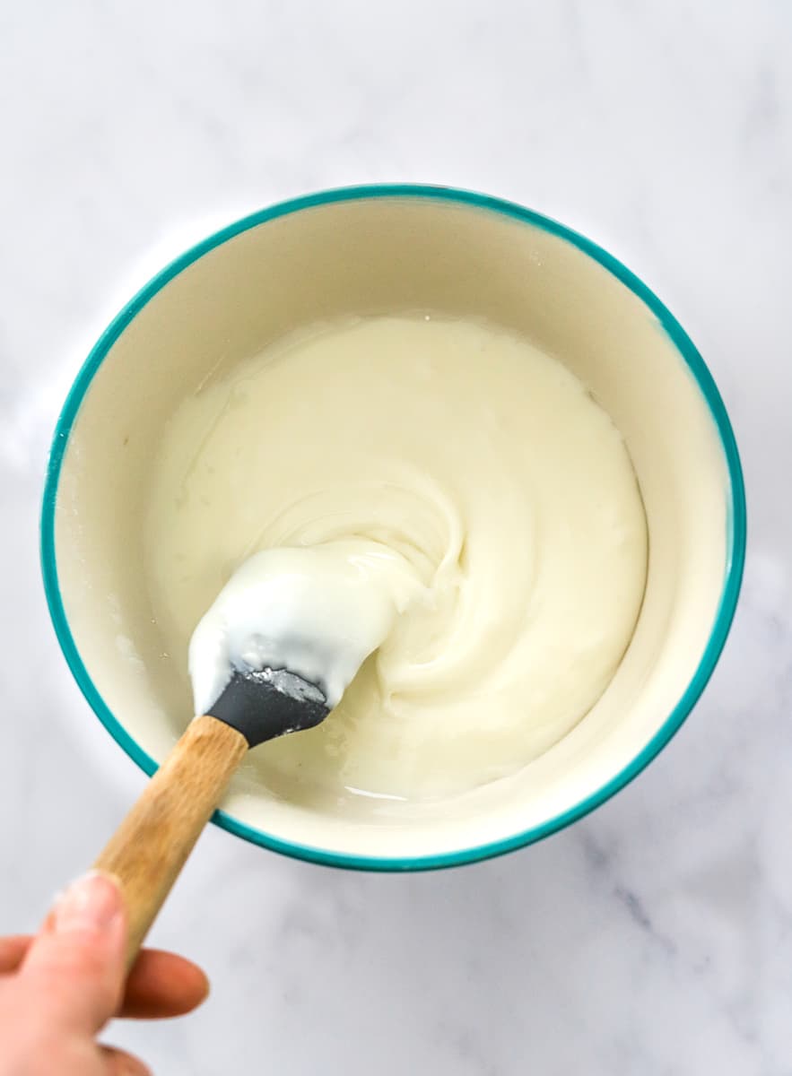 Hand mixing white frosting with a grey rubber spatula in a bowl with a blue rim.