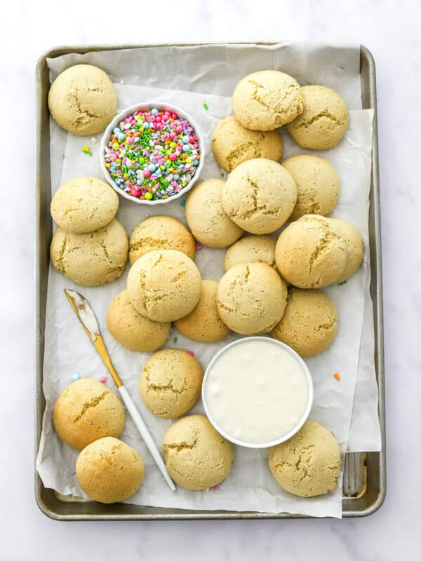 Silver tray lined with white paper with unbaked round light coloured cookies on it with a bowl of coloured sprinkles and a bowl of white frosting and a knife on the tray with the cookies.