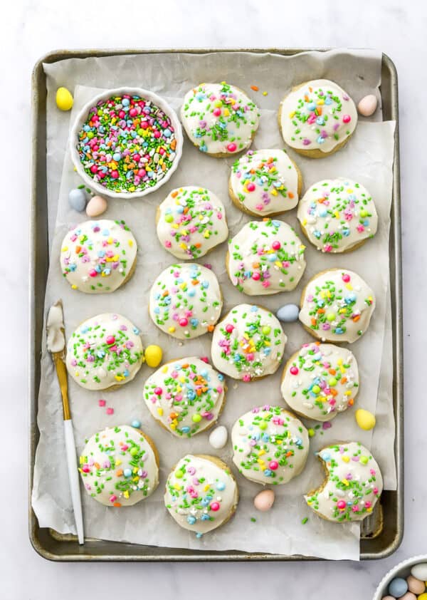Iced round easter cookies with colourful sprinkles on them on a lined tray with a bowl of more sprinkles and a white knife on the tray with the cookies.
