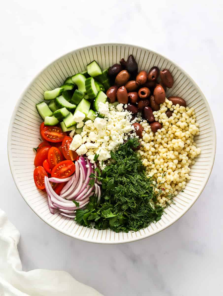 Chopped tomatoes, cucumber, dill, parsley, red onion, olives feta and cooked Israeli couscous in a round white bowl with a white linen in front of it.