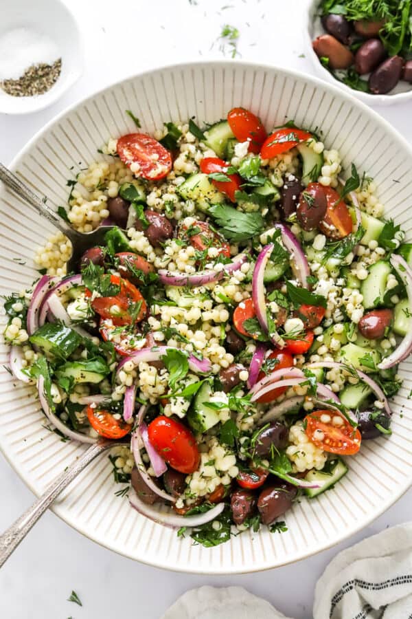 Mediterranean salad with tomatoes, olive, and feta cheese toss with cooked couscous couscous in a white salad bowl with more salad ingredients around it.