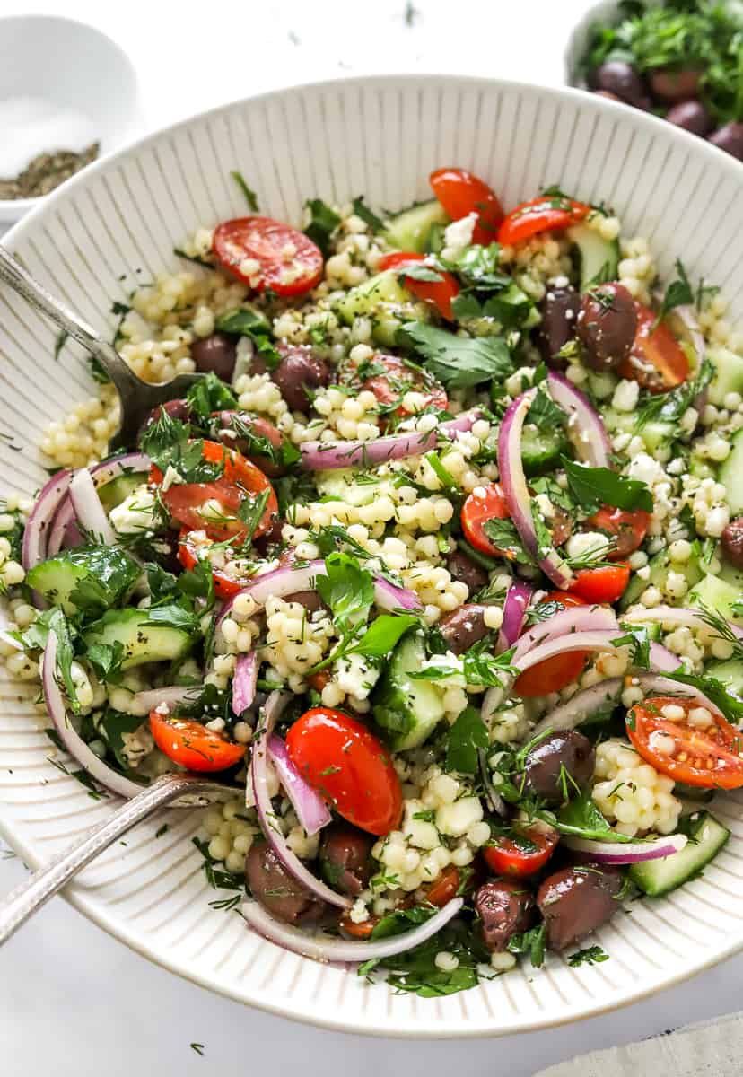 Israeli couscous salad in a white lined serving bowl with green and red veggies and herbs on it with serving spoons in the bowl.